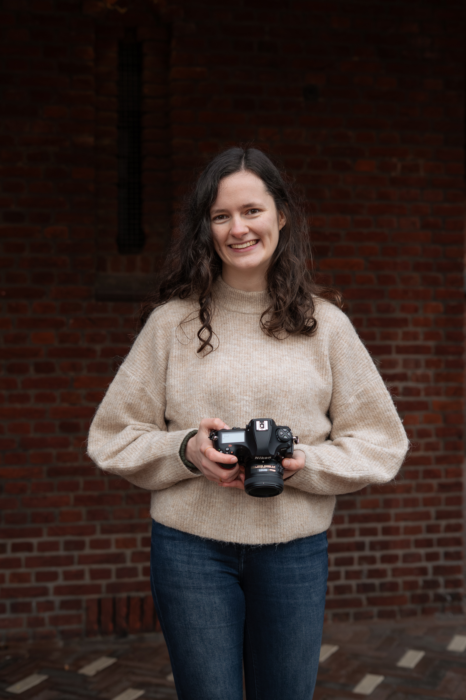 Natürliches Porträtfoto einer Fotografin in Ahaus, die eine Kamera in der Hand hält und lächelt, vor einer Backsteinwand eines historischen Gebäudes.