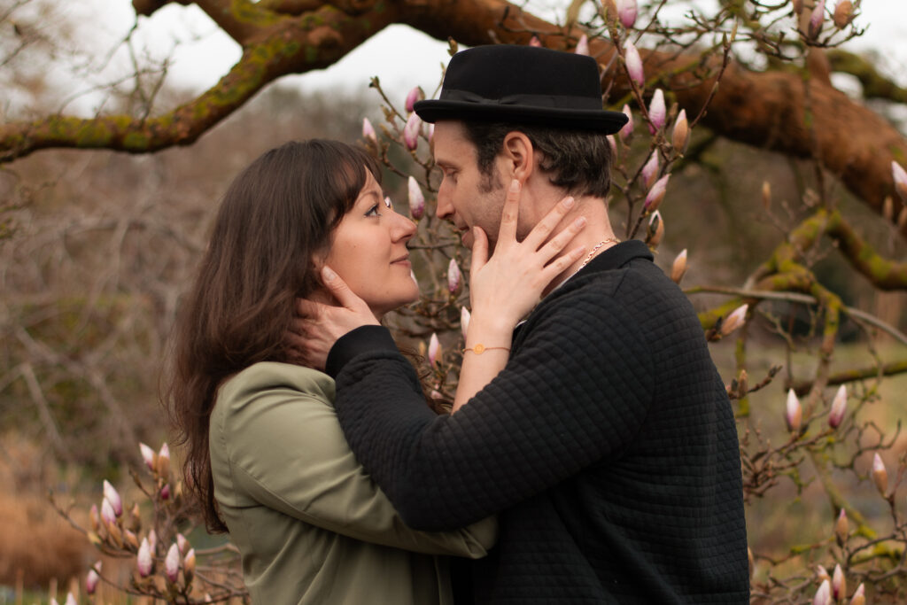 Paarfoto eines Paars aus Frau und Mann vor einem Magnolienbaum mit noch geschlossenen Blüten. Das Paar schaut sich an, lächelt leicht und legt jeweils eine Hand an das Gesicht der oder des anderen.