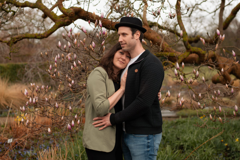Ein Paar aus Frau und Mann auf einem Paarfoto, auf dem die Frau ihren Kopf an die Brust des Mannes lehnt und leicht lächelt. Das Paar steht vor einem Magnolienbaum mit noch geschlossenen Blüten.