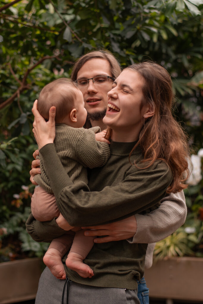 Familienfoto mit einer Frau, einem Mann und einem Kleinkind in einem Gewächshaus. Die Frau hält das Kind auf den Arm. Der Mann hat seine Hände um die beiden gelegt. Der Mann schaut das Kind an und die Frau lacht.