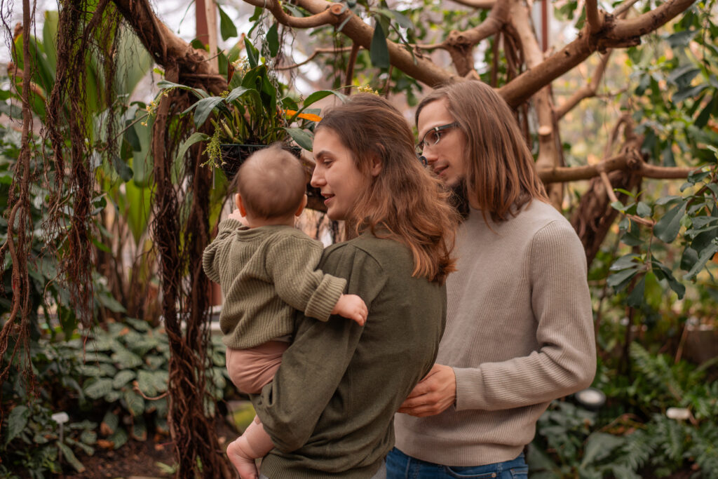 Familienfoto mit einer Frau, einem Mann und einem Kleinkind in einem Gewächshaus. Die Frau hält das Kind auf dem Arm, während sie und der Mann es anschauen.