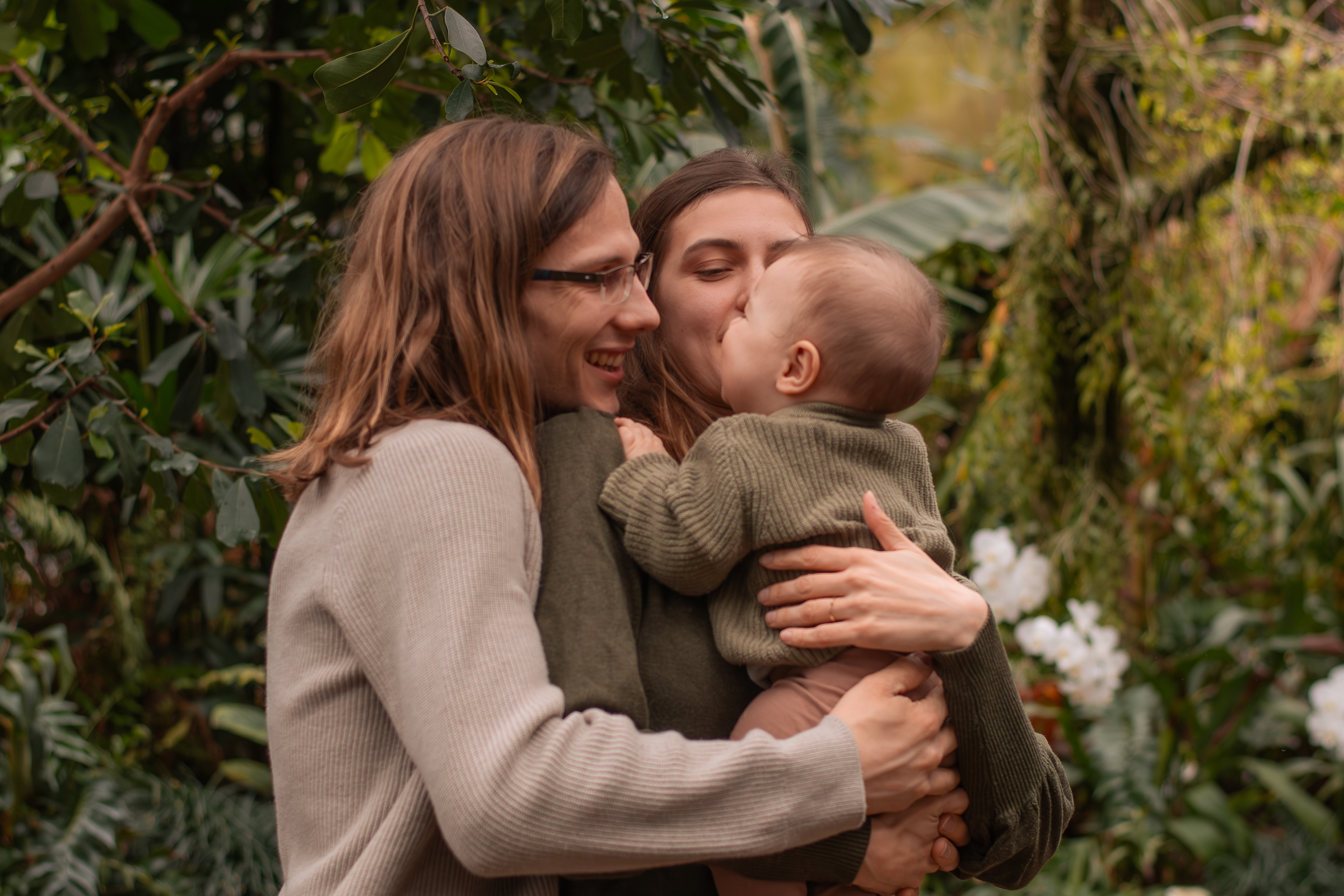 Natürliches Familienfoto aus einem Familienshooting mit einer Frau, einem Mann und einem kleinen Kind. Die Frau hält das Kind auf dem Arm, der Mann umarmt beide von hinten und alle lachen.