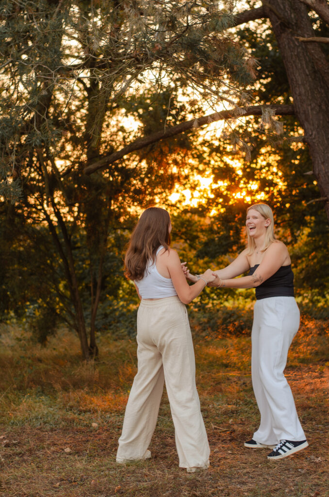 Natürliches Foto aus einem Freundinnenshooting mit zwei Freundinnen. Sie stehen sich in einem Park vor der untergehenden Sonne gegenüber, halten sich an den Händen und lachen.