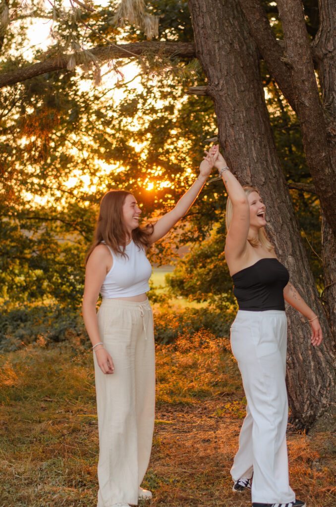 Freundinnenshooting mit zwei Freundinnen in einem Park. Sie halten sich jeweils mit einer Hand über ihren Köpfen fest wie bei einer Drehung beim Tanzen und lachen.