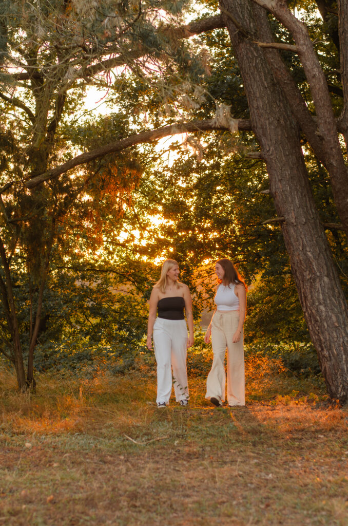 Freundinnenshooting mit zwei Freundinnen, die über eine Wiese laufen und sich anschauen. Hinter ihnen scheint die untergehende Sonne durch die Bäume.