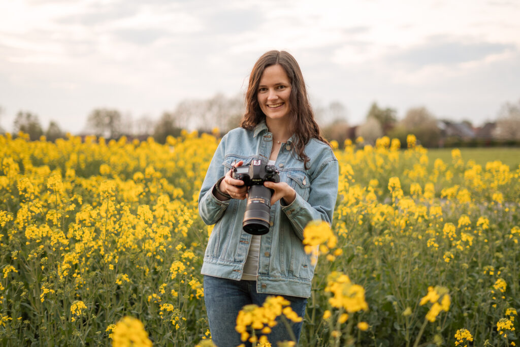 Natürliches Porträt einer Fotografin aus Ahaus im Münsterland mit einer Kamera in den Händen in einem Rapsfeld mit gelben Blüten.