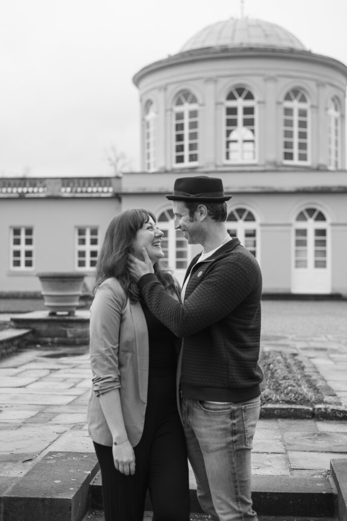 Schwarz-Weiß-Foto aus einem Paarshooting mit einer Frau und einem Mann, das sich anschaut und lacht, vor einem historischen Gebäude mit oben abgerundeten Fenstern.