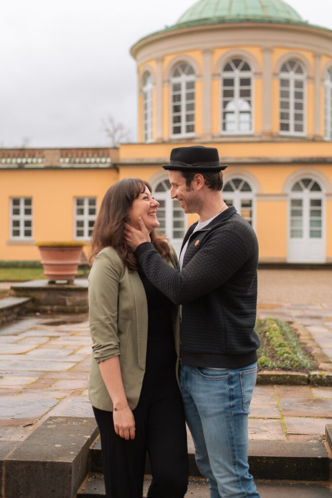 Paarfoto aus einem Paarshooting mit einem Mann und einer Frau vor einem historischen, gelben Gebäude mit oben abgerundeten Fenstern. Das Paar schaut sich in die Augen und lacht, der Mann hat seine linke Hand an den Hals der Frau gelegt.