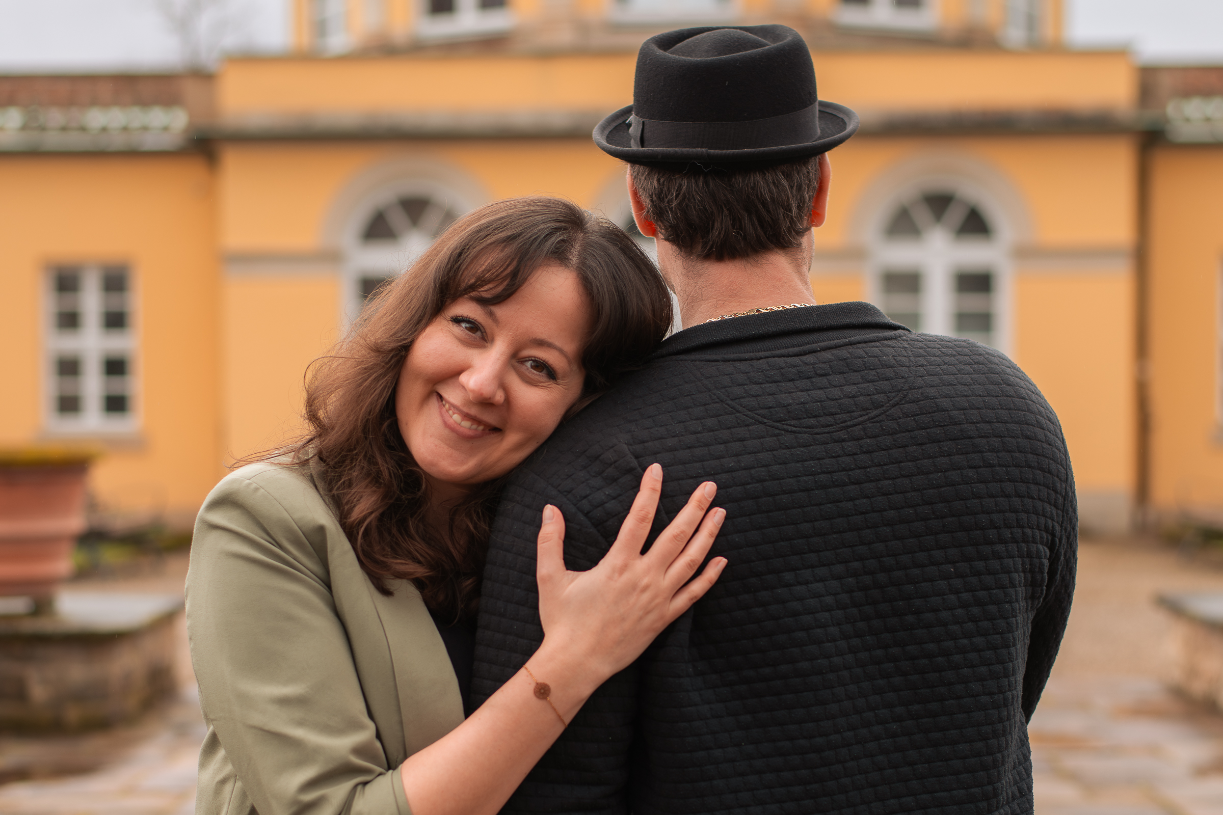 Paarfoto mit einer Frau und einem Mann vor einem historischen Gebäude mit oben abgerundeten Fenstern. Die Frau legt ihren Kopf auf die Schulter des Mannes und lächelt in die Kamera.