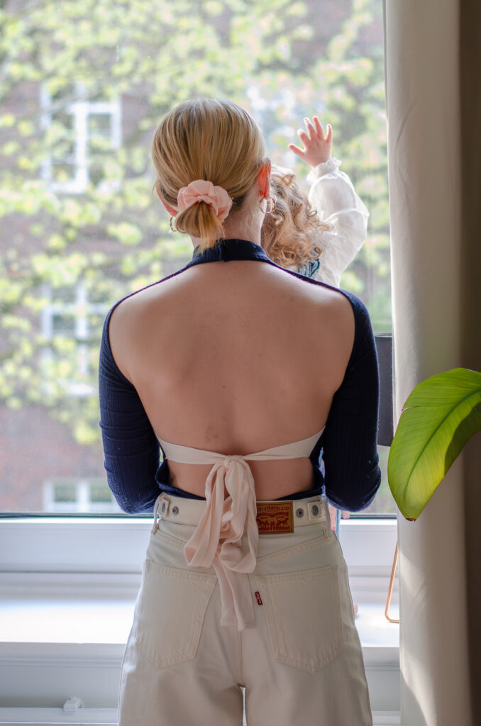 Foto aus einem Familienshooting mit einer Mutter und ihrer Tochter. Sie stehen am Fenster und die Tochter hebt ihre rechte Hand am Fenster hoch. Die Mutter hält die Tochter fest. Beide kehren der Kamera den Rücken zu.