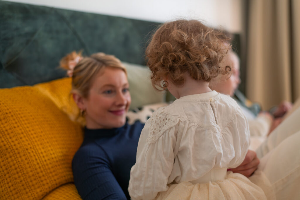 Familienshooting mit einer Mutter und ihrer Tochter. Sie sitzen auf dem Bett, die Tochter auf dem Schoß ihrer Mutter mit dem Rücken zur Kamera. Die Mutter schaut ihre Tochter an und lächelt.