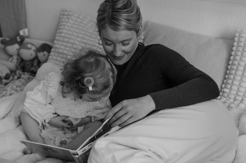 Schwarz-Weiß-Foto aus einem Familienshooting mit einer Mutter und ihrer kleinen Tochter. Sie sitzen zusammen im Bett und schauen sich ein Kinderbuch an.
