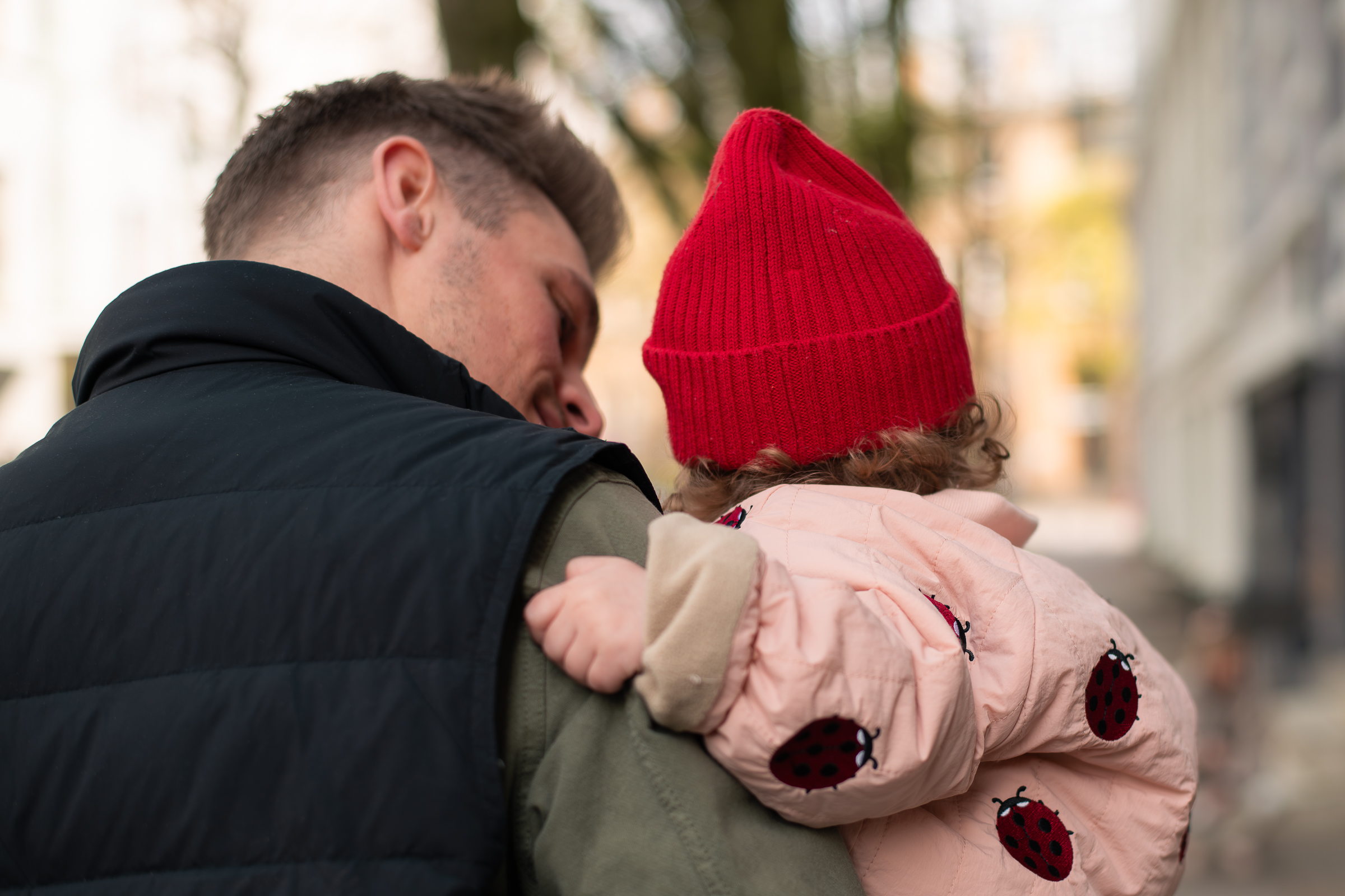 Familienfoto mit einem Vater und seiner kleinen Tochter. Der Vater hat seine Tochter auf dem Arm, schaut sie an und lächelt. Beide kehren der Kamera den Rücken zu.