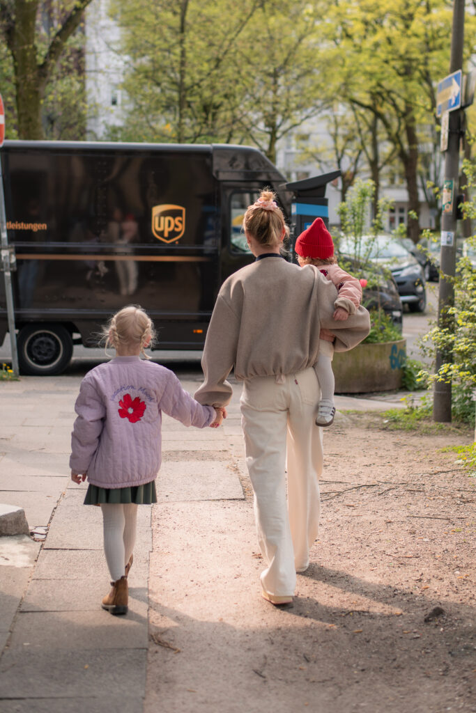 Natürliches Foto aus einem Familienshooting mit einer Mutter und ihren beiden Töchtern beim Spaziergang. Sie hält ihre ältere Tochter an der linken Hand und trägt die jüngere Tochter auf dem rechten Arm.
