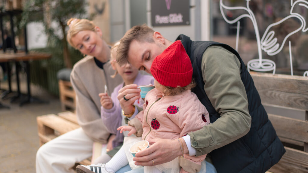 Familienfoto mit Mutter, Vater und zwei Töchtern beim Eis essen. Sie sitzen auf einer Bank aus Paletten. Der Vater hat die kleinste Tochter auf dem Schoß und füttert sie. Links neben ihnen sitzen die ältere Tochter und die Mutter, die die jüngere Tochter anschauen.
