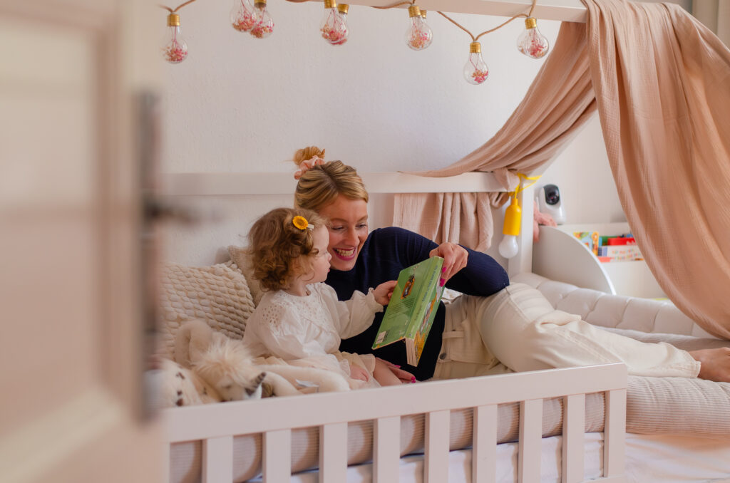 Foto aus einem Familienshooting mit einer Mutter und ihrer kleinen Tochter. Sie sitzen zusammen im Bett und schauen sich ein Kinderbuch an.