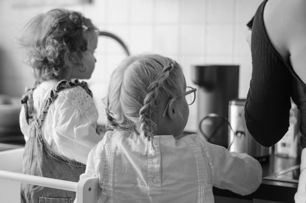 Familienfoto mit zwei Töchtern in der Küche. Sie schauen ihrer Mutter dabei zu, wie sie etwas zubereitet. Alle kehren der Kamera den Rücken zu.